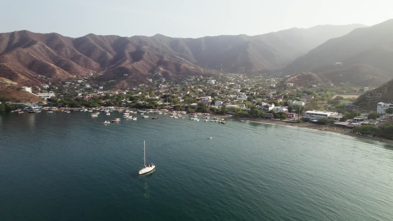 Stunning aerial dolly zoom shot over the picturesque bay of Taganga, Santa Marta. The camera pulls back from a small sailboat, revealing the Caribbean coastline and the surrounding arid Sierra Nevada