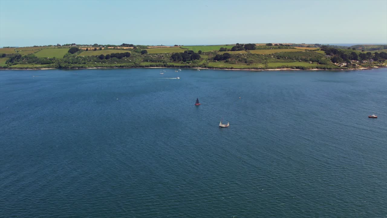 Aerial footage of boats sailing on a calm bay with a scenic coastline of green fields and trees in the background. Smooth lateral camera movement.