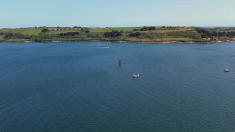 aerial footage of boats sailing on a calm bay with a scenic coastline of green fields and trees in the background. smooth lateral camera movement.