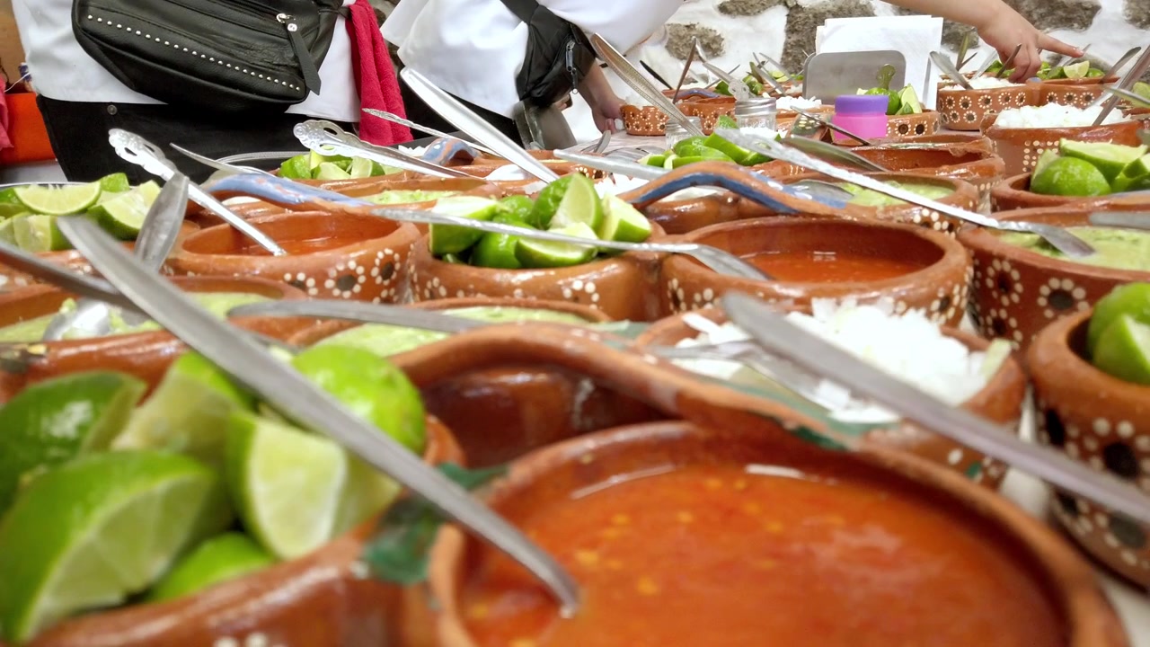 Buffet di cibo messicano con ciotole di argilla decorate piene di salse, spicchi di lime fresco, utensili da portata e persone che preparano piatti in un servizio di ristorazione in Messico.
