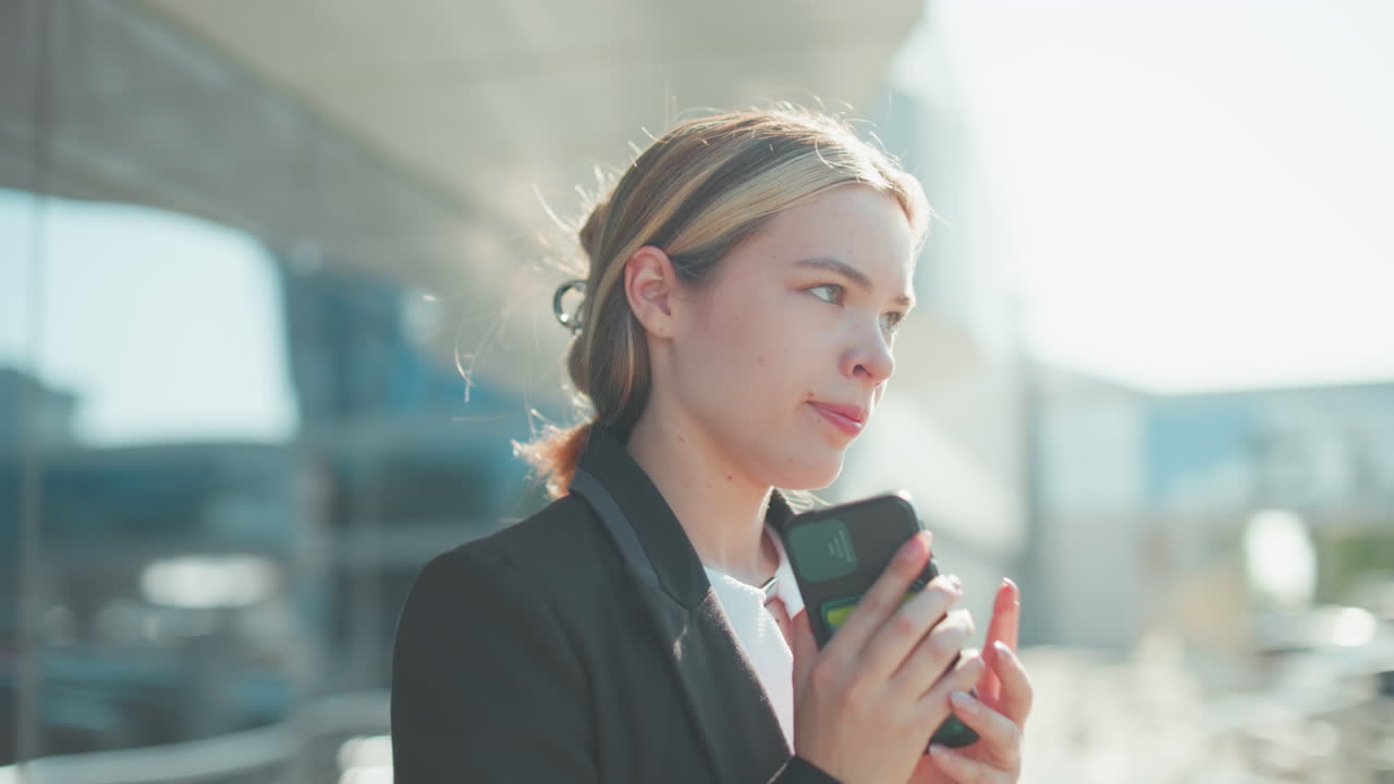 Frustrated woman during phone call lowers phone briefly, continues conversation after work while exiting office building, dressed in formal attire, glass door reflects cityscape