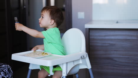 lovely little toddler boy sitting at white feeding table. kid eats with fork in left hand then changes to right hand with mom's help.