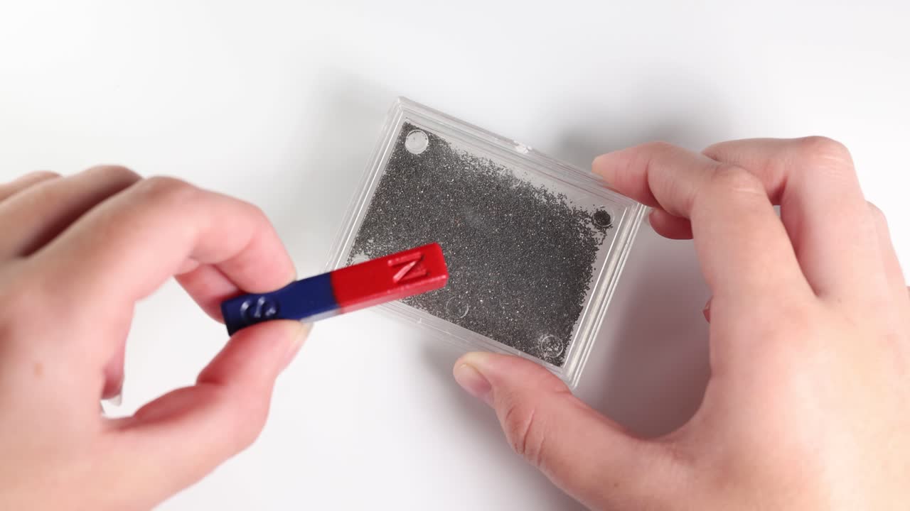 Hands manipulate a magnet over iron filings, illustrating magnetic field patterns. Bright lighting enhances visibility of the scientific demonstration