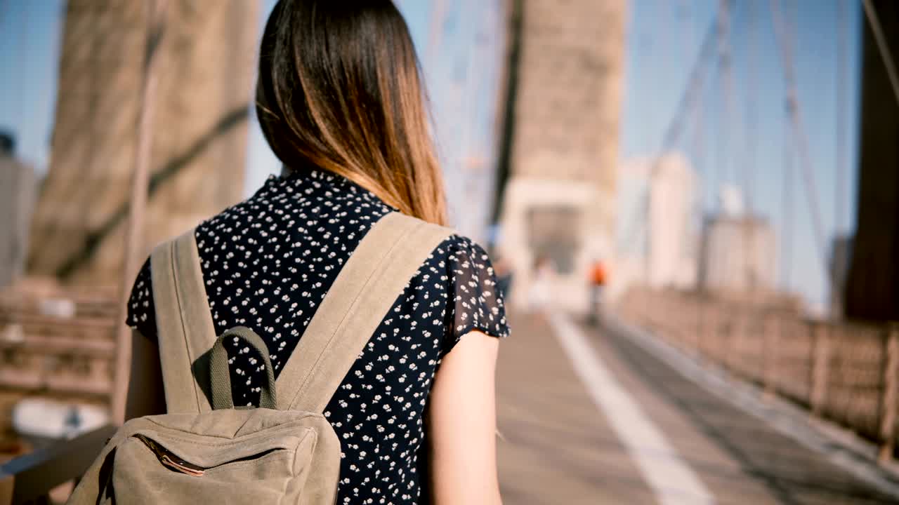 Camera follows female tourist with backpack and dark golden hair walking along Brooklyn Bridge on a clear sunny day 4K