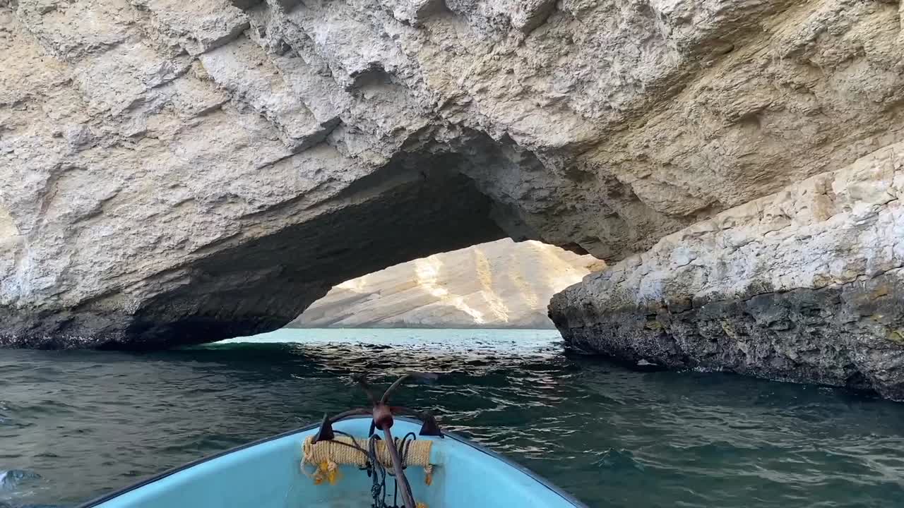 A ship with an anchor slowly passes under a natural stone arch near Qantab beach in Muscat, Oman