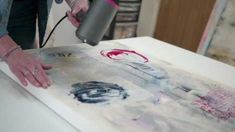 Female-artist-using-a-hair-dryer-to-dry-a-canvas,-smoothing-out-material,-stood-in-a-home-studio