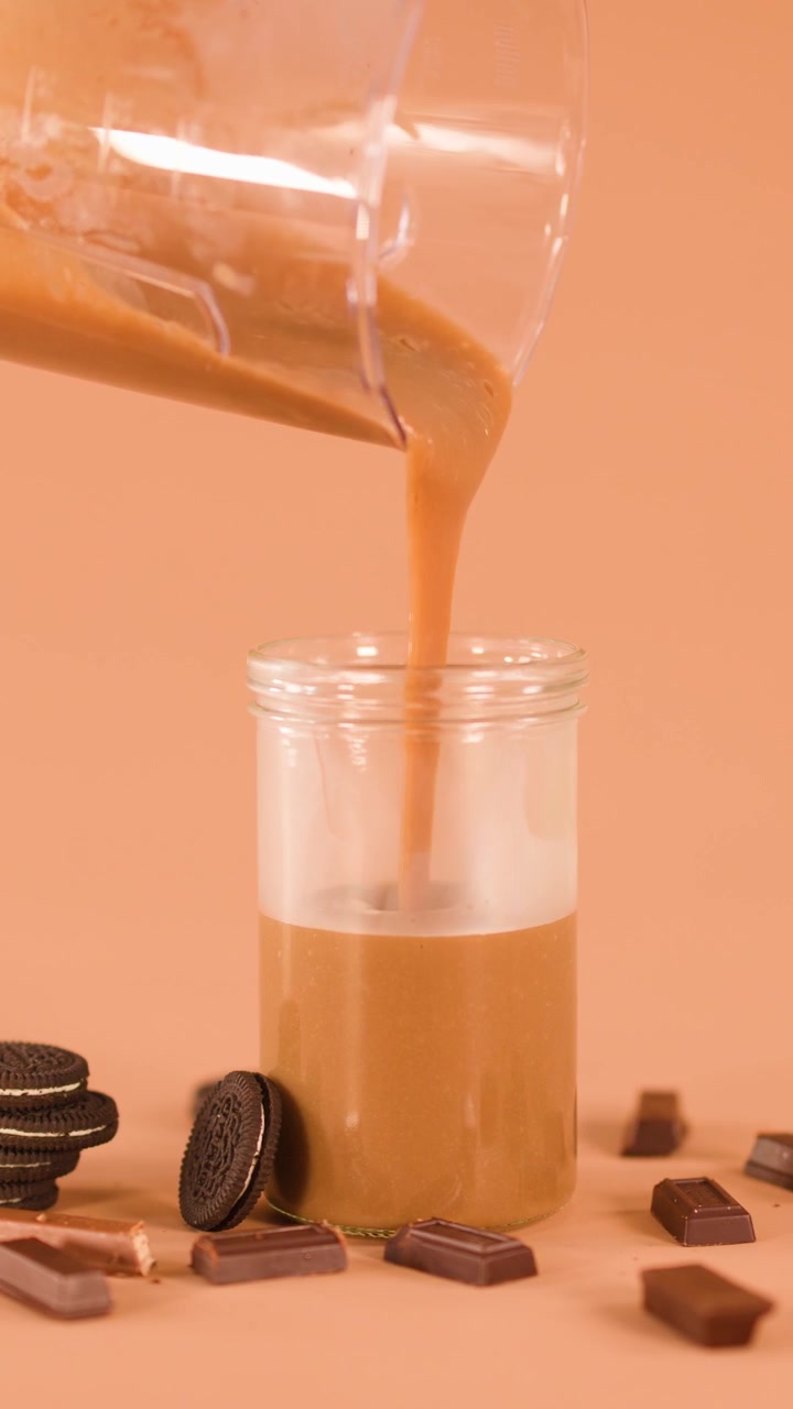 Bevanda al cioccolato versata dal frullatore in un barattolo di vetro, circondata da biscotti e barrette di cioccolato.
