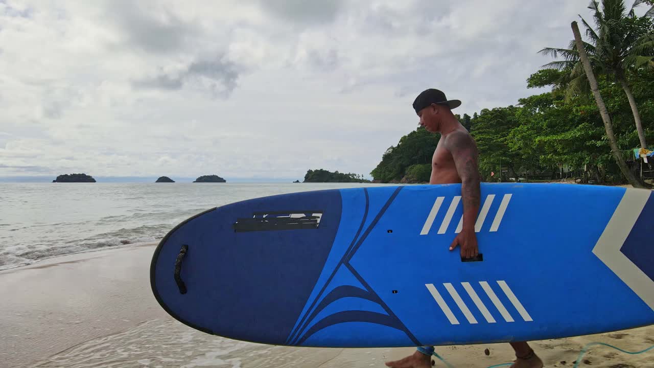 hombre asiático caminando con una tabla de remo en una playa hacia el océano con islas y costas en tailandia