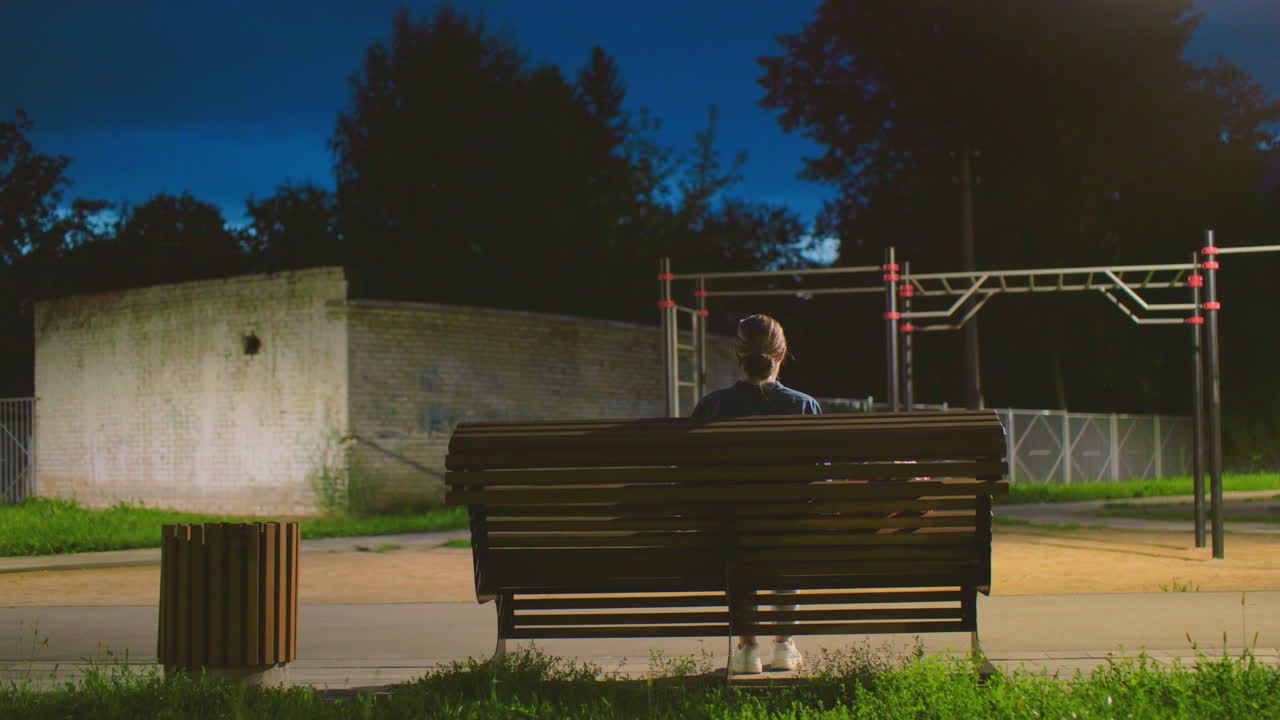 vista trasera de una mujer sentada en un banco al aire libre durante la noche, frente a una estructura de patio de recreo bajo una luz tenue, con un entorno sombreado, una pared en el fondo y un cielo azul profundo