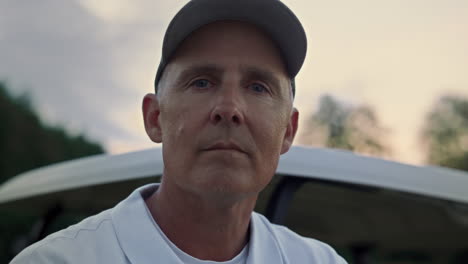 pensive senior golfer standing at golf cart. old guy looking camera at sunset.