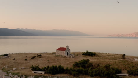 cinematic drone shot revealing a tiny island with a church in the middle of the sea, sunrise, blue hour, seagulls flying around, calm water, peaceful clear view, chapel, novi vinodolski, croatia