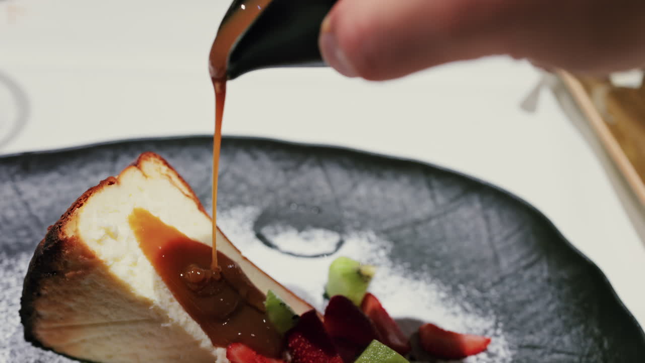 Close up of caramel sauce being poured over a slice of cheesecake served with fresh strawberries and kiwi, garnished with powdered sugar on a plate at a restaurant
