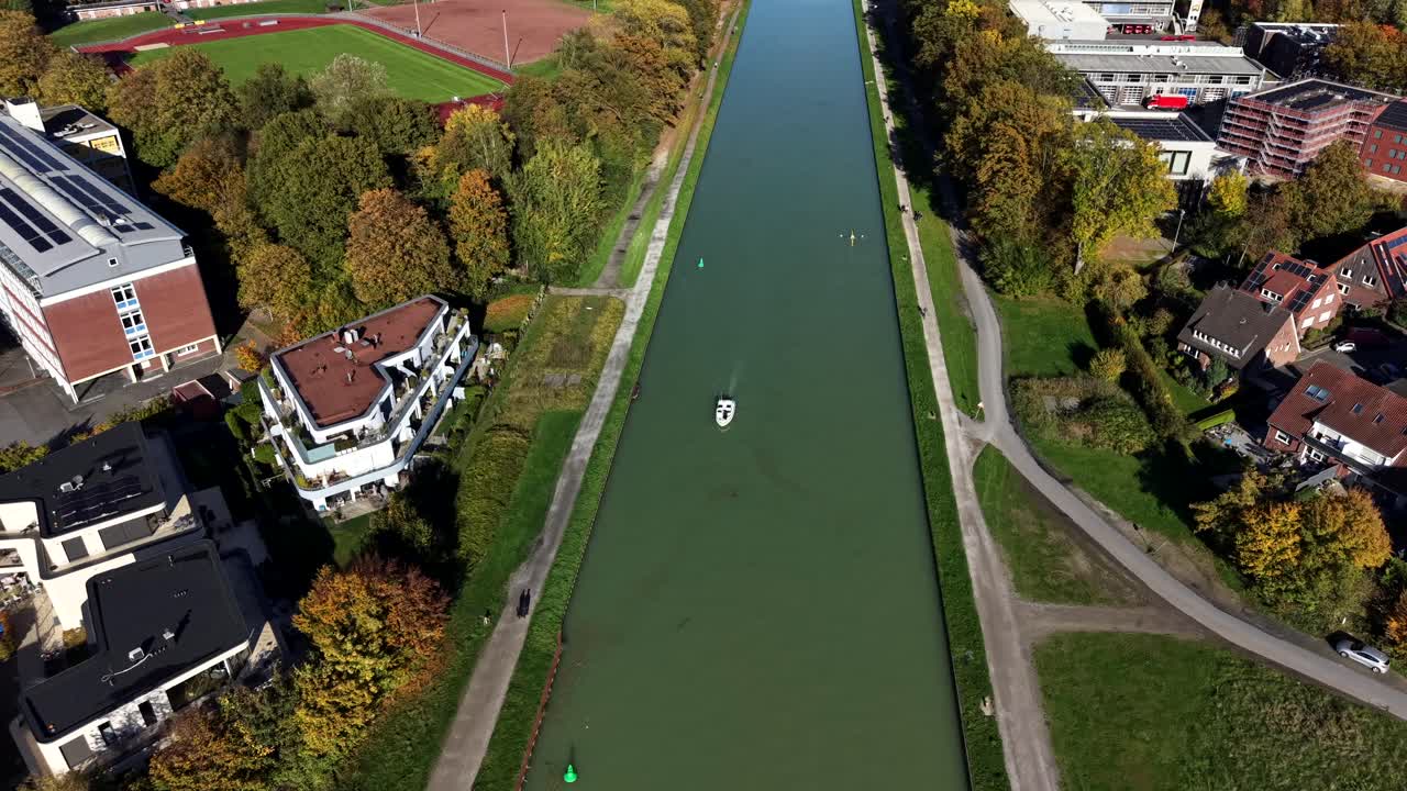 Aerial flyover green river in luxury suburb neighborhood near soccer field in autumn season. Sunny day day in german town at fall. Birds eye shot. Cruising boat on water