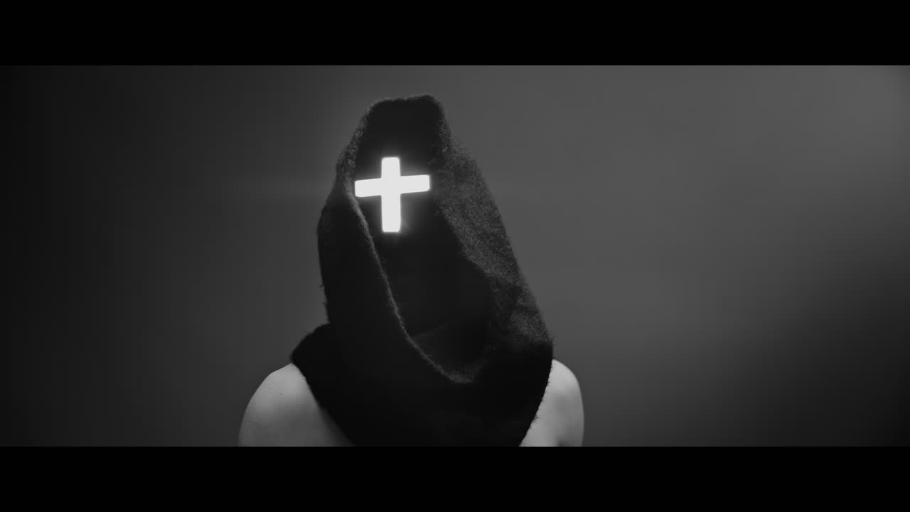 Masked and hooded man with cross of light glowing on his face looks in the camera