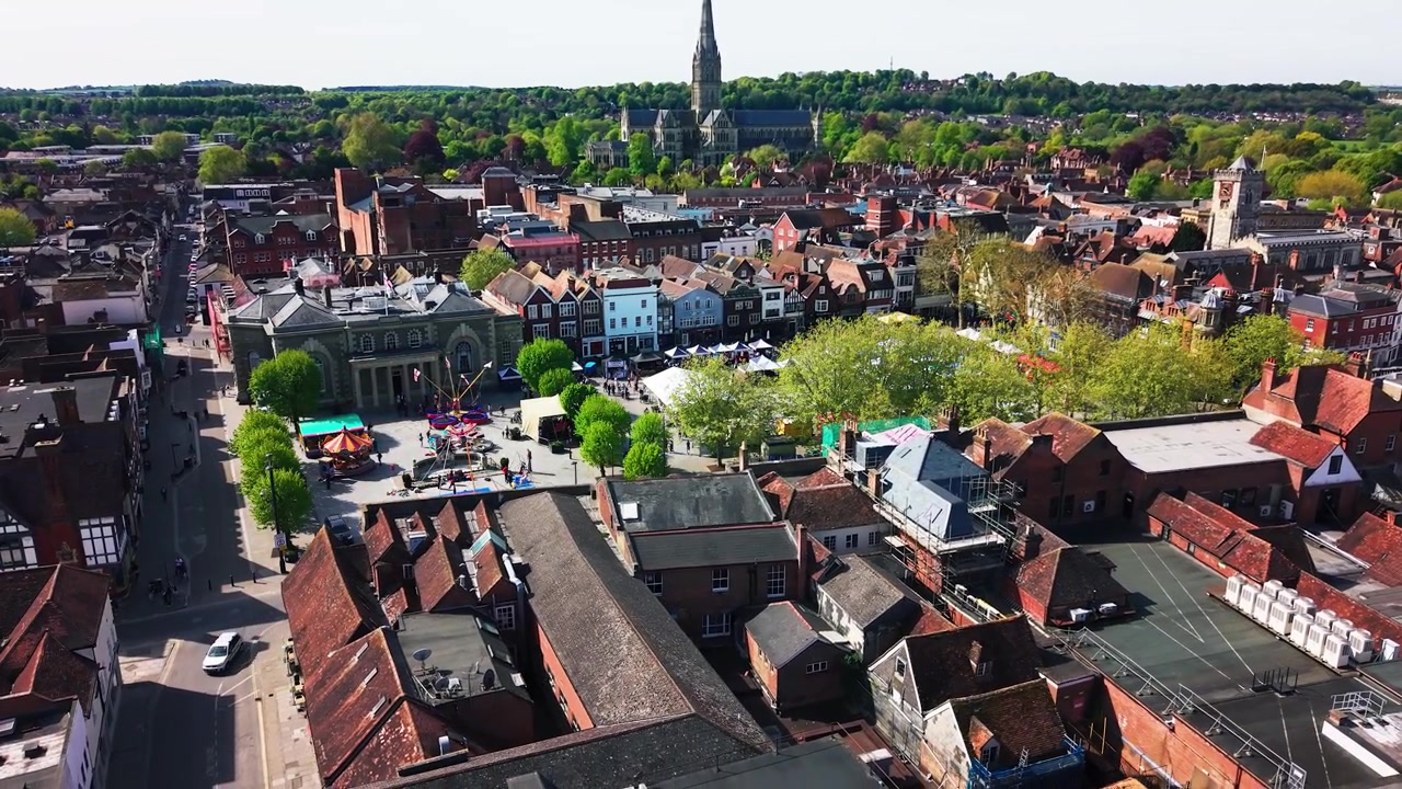 Wide aerial drone footage of Salisbury Market Square during a St George’s Day event, showing the Guildhall, event stalls, fairground rides, crowds, trees, rooftops, roads, and historic city centre