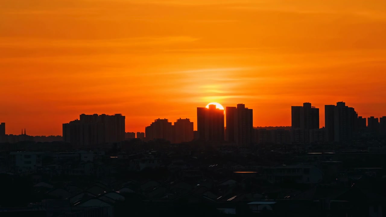 Dark urban buildings stand in silhouette along the horizon as the sun glows near the skyline at dusk. Warm backlight fills the sky with calm evening atmosphere and a dramatic city view