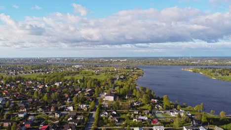 High aerial of suburban rooftops and green lawns in peaceful spring neighborhood, tilt up along river and road to clouds