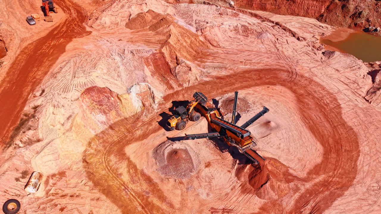 Madrid, Spain, 8 August 2025: Birds eye view of heavy machinery in red clay quarry. Overhead shot of a mobile screening plant and wheel loader processing raw materials in an open pit mine