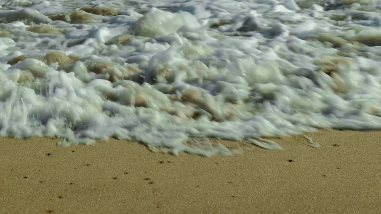 A slow-motion shot captures waves gently breaking over golden sand, creating soft foam as they roll in, showcasing the beauty of the shoreline.