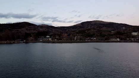 Crepúsculo-Sobre-Un-Lago-Tranquilo-Con-Un-Paisaje-Montañoso-Y-Casas-Al-Fondo,-Ambiente-Sereno