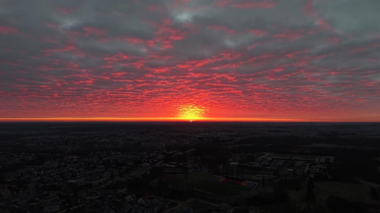 Atemberaubendes Luftbildmaterial fängt einen lebendigen Sonnenaufgang ein, der den Himmel in Rottönen über einer weitläufigen Stadtlandschaft malt. Die Wolkenstruktur verleiht der ruhigen und friedlichen Morgensicht Dramatik.