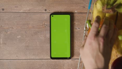 overhead studio shot of person picking from basic food items in supermarket wire shopping basket next to green screen mobile phone