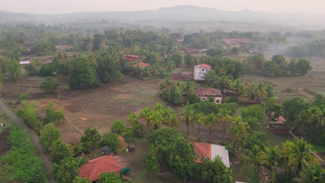 a stunning drone shot of avalegaon, kudal village in konkan, maharashtra, india showcasing a serene traditional houses, lush green trees, vast fields and majestic mountains under a clear pink sky