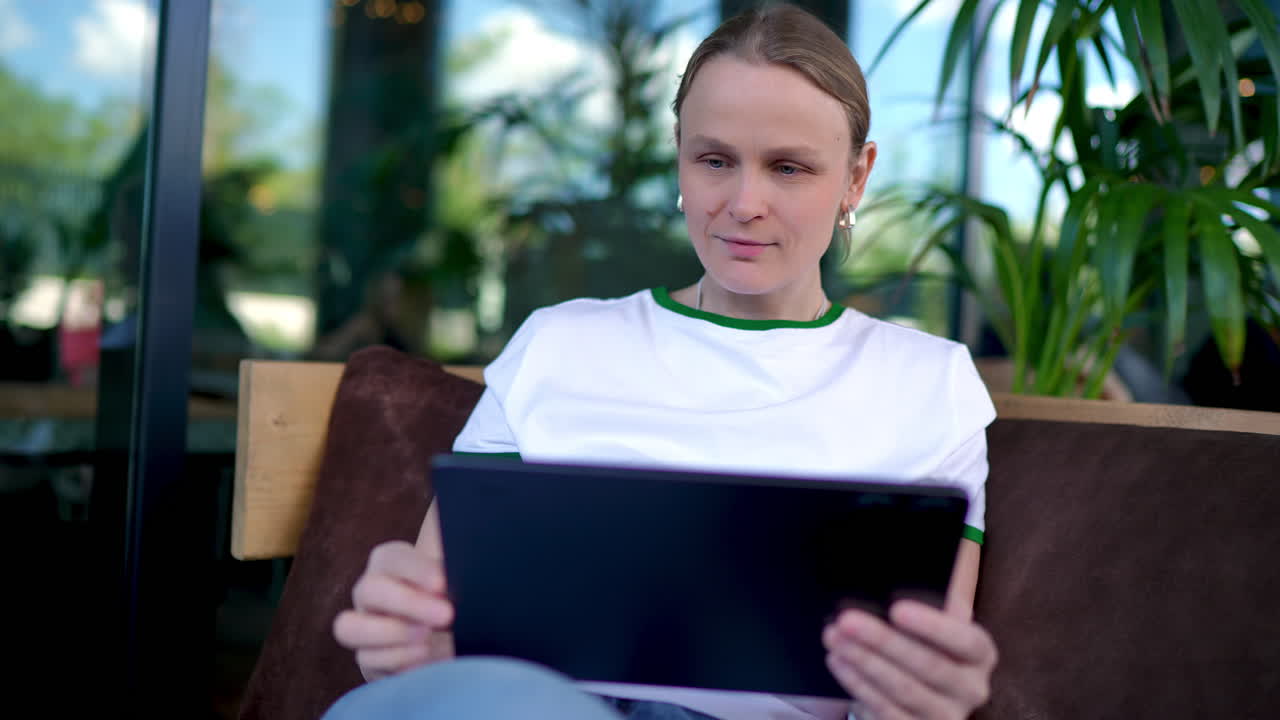 Young woman working remotely, sitting comfortably at outdoor cafe, browsing tablet while enjoying peaceful moment in stylish urban setting with greenery in background