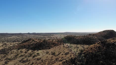 Hills-covered-with-grass,-red-sand,-bushes,-trees-and-rocks-are-captured-by-a-camera-moving-over-a-large-African-savanna