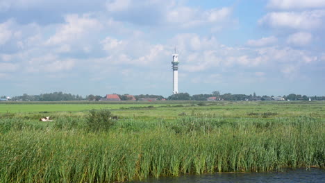 Paisaje-Holandés-Plano-Con-Una-Alta-Torre-De-Telecomunicaciones,-Toma-En-Movimiento-Desde-Un-Barco