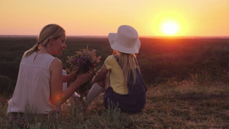 mom and little daughter are sitting on a hill admiring the sunset in the hands of wild flowers summe