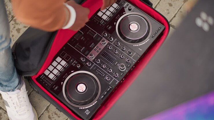 Controller DJ estratto per un set di strada, artista con giacca marrone solleva il controller da una borsa con profili rossi accanto a un altoparlante colorato, le mani regolano il deck e i cavi sul marciapiede, sneakers e jeans strappati.