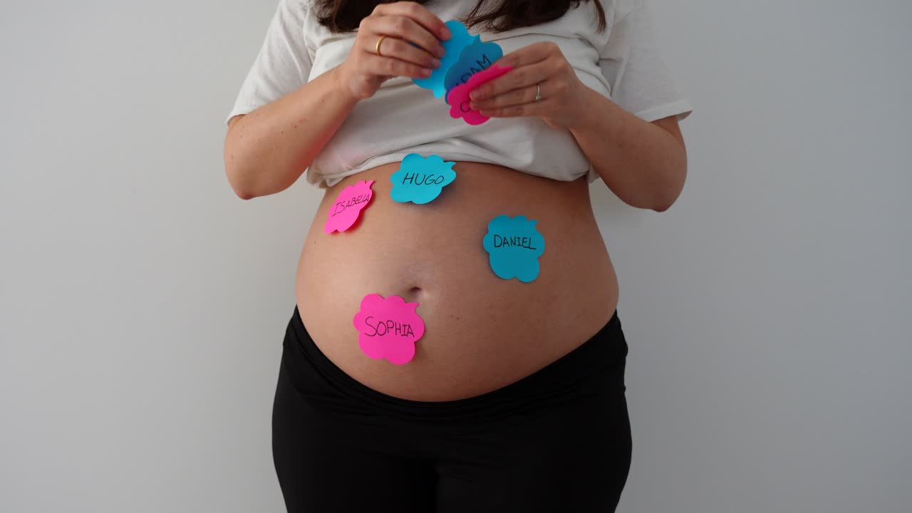 Pregnant woman places pink and blue baby name notes on her belly at home, white background