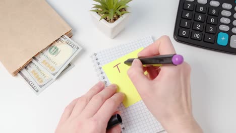 las manos escriben una nota de día de impuestos en papel adhesivo amarillo. el concepto de impuestos, dinero y una calculadora se encuentran a su lado.