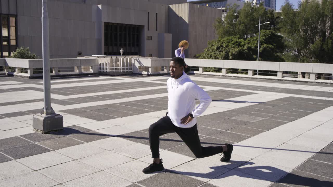 African American man in hoodie stretching, lunging on roof warming while woman phoning