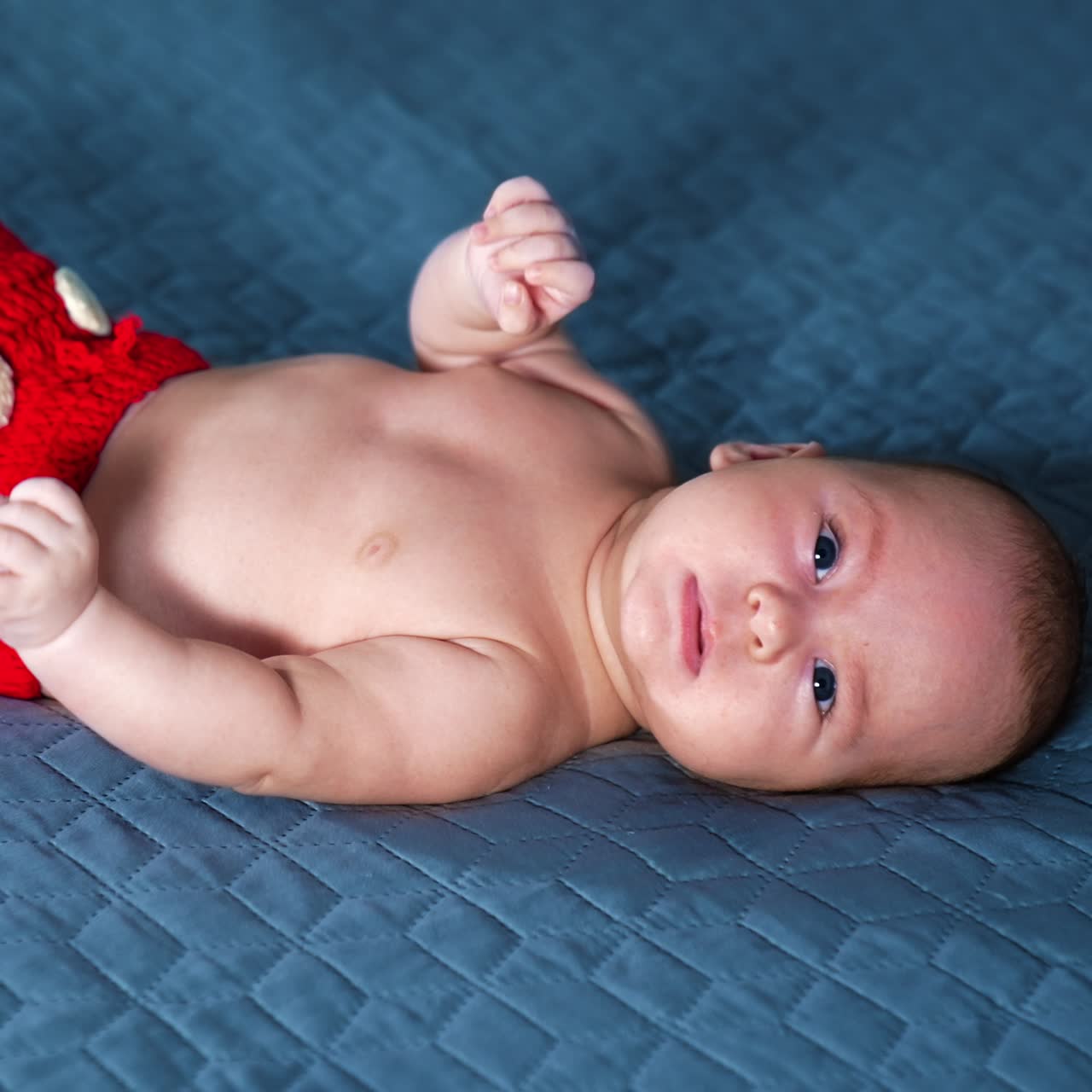Happy little baby in red shorts with white buttons beats his feet and hands. Active kid in costume at grey backdrop