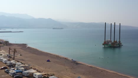 HaDatiyim-Beach-Line-With-Many-Camping-Cars-Parked-in-Eilat-city,-Aerial-View-of-Offshore-Self-elevating-Platform-with-Massive-Columns