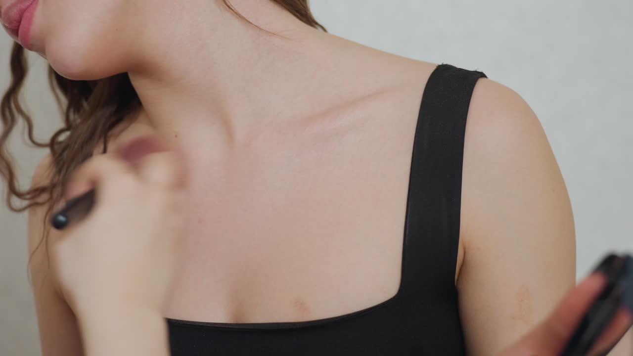 White Woman Receiving Clavicle Highlight, Focused Brushwork Along Collarbone And Shoulder, Subtle Shimmer Application For Photoshoot Glow, Sleek Black Top, Professional Touch, Soft Neutral Backdrop