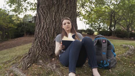 female college student takes notebook out of backpack to write in journal under a tree at campus park