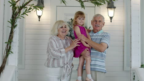 Happy-senior-grandfather-and-grandmother-couple-holding-granddaughter-in-hands-in-porch-at-home