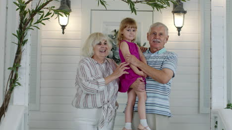 happy senior grandfather and grandmother couple holding granddaughter in hands in porch at home