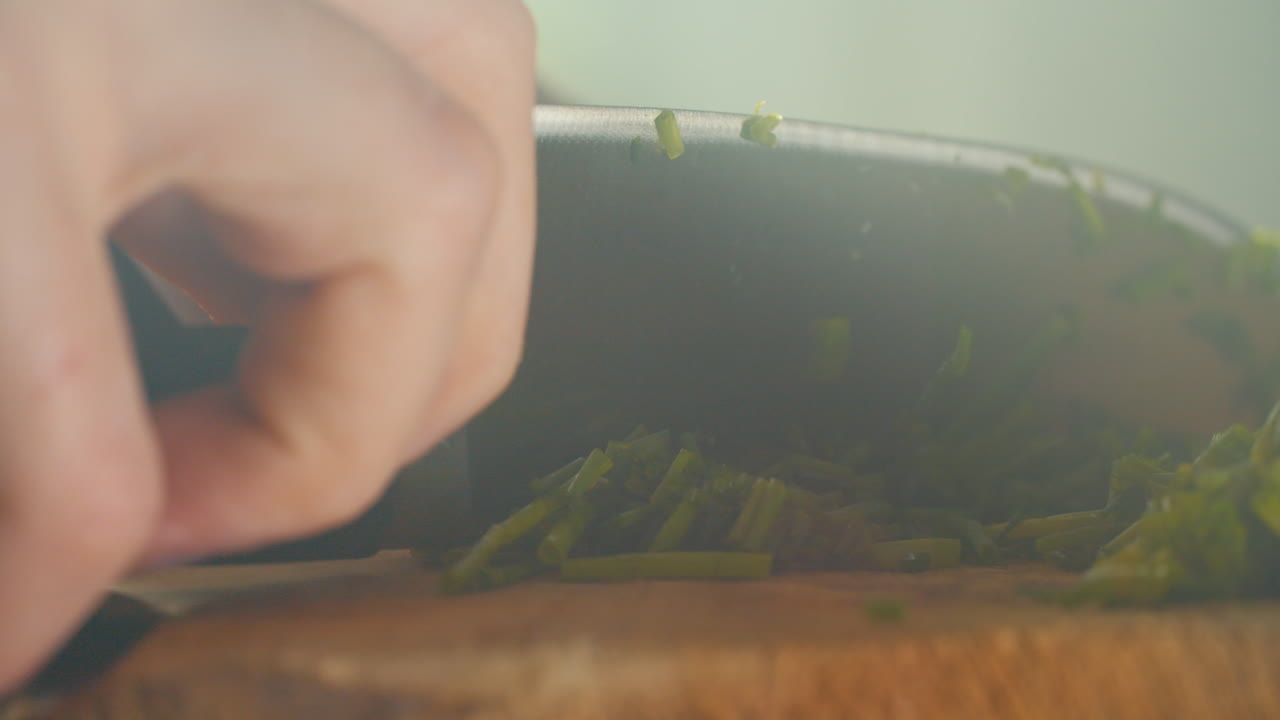 Primo piano di una mano maschile che taglia l'erba cipollina su un tagliere di legno in un'atmosfera di cucina fumosa, con bellissimi raggi di sole.