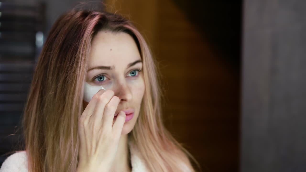 Blond kvinne i morgenkåpe fjerner hvite øyepads mens hun ser seg i speilet