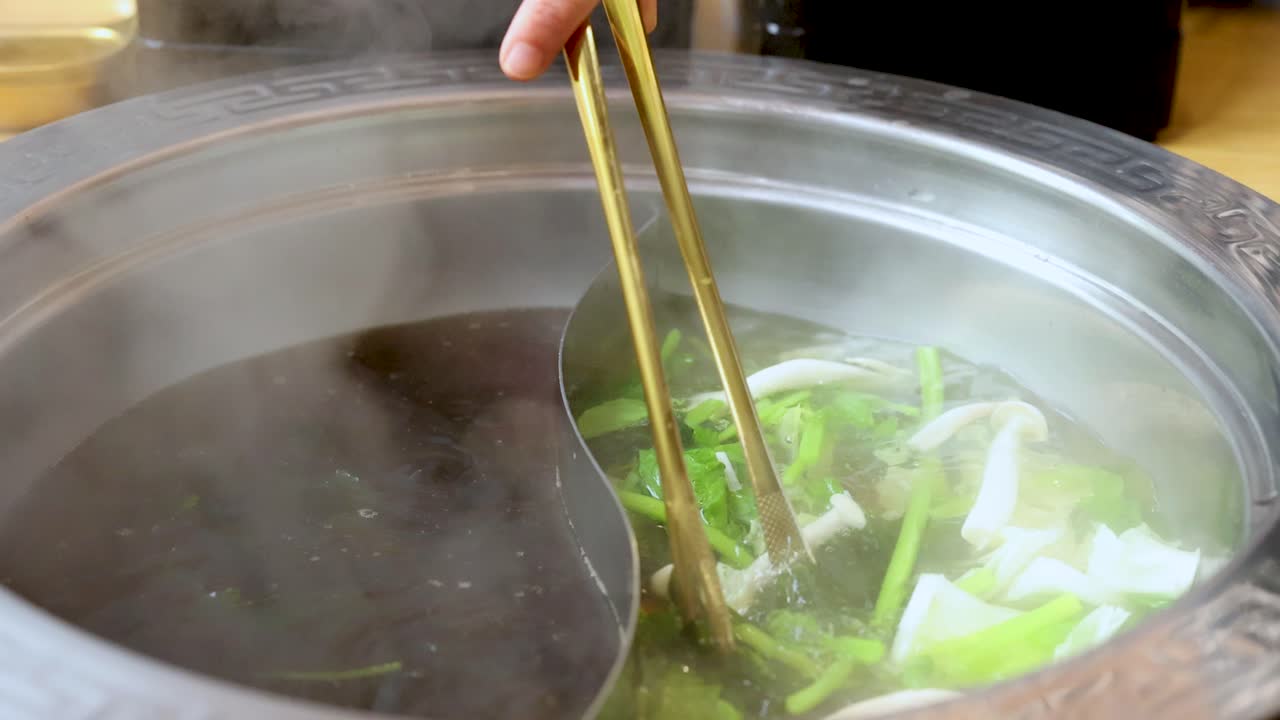 Hand stirs leafy greens and mushrooms in steaming dual broth hotpot under bright indoor lighting
