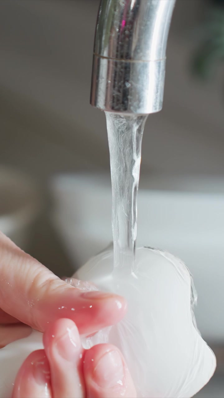 Close up of hand holding. Object becomes wet and shiny as hand moves it around with water consistently. Background shows blurred sink setting with white bowls showing hand