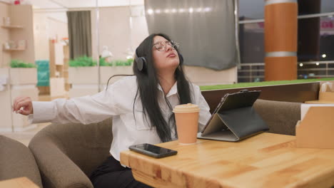 side view of young woman in headphones working on tablet at cafe desk while stretching herself slightly, appearing focused and relaxed with coffee on table and warm indoor ambiance around her