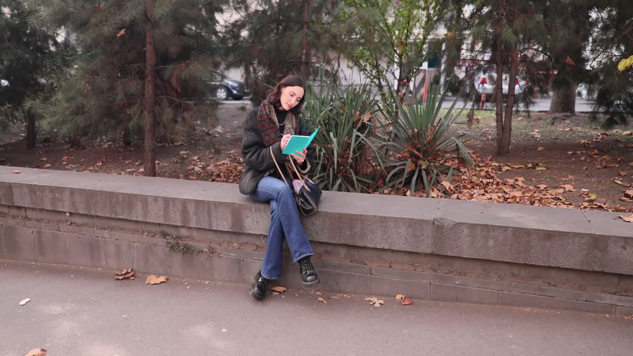Young woman writing or reading in a notebook while sitting on a wall in a park