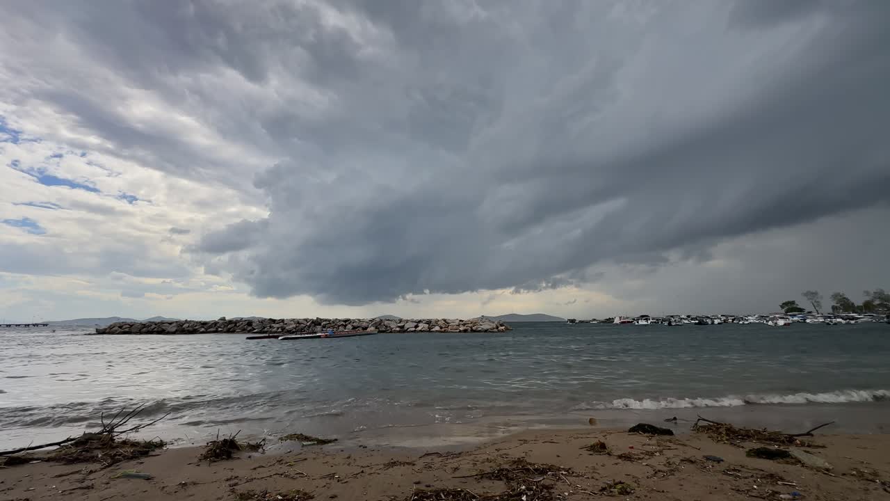 Dramatic dark storm clouds gather over a rough sea. Waves crash on the shore near a stone breakwater and a marina with boats on a gloomy autumn day