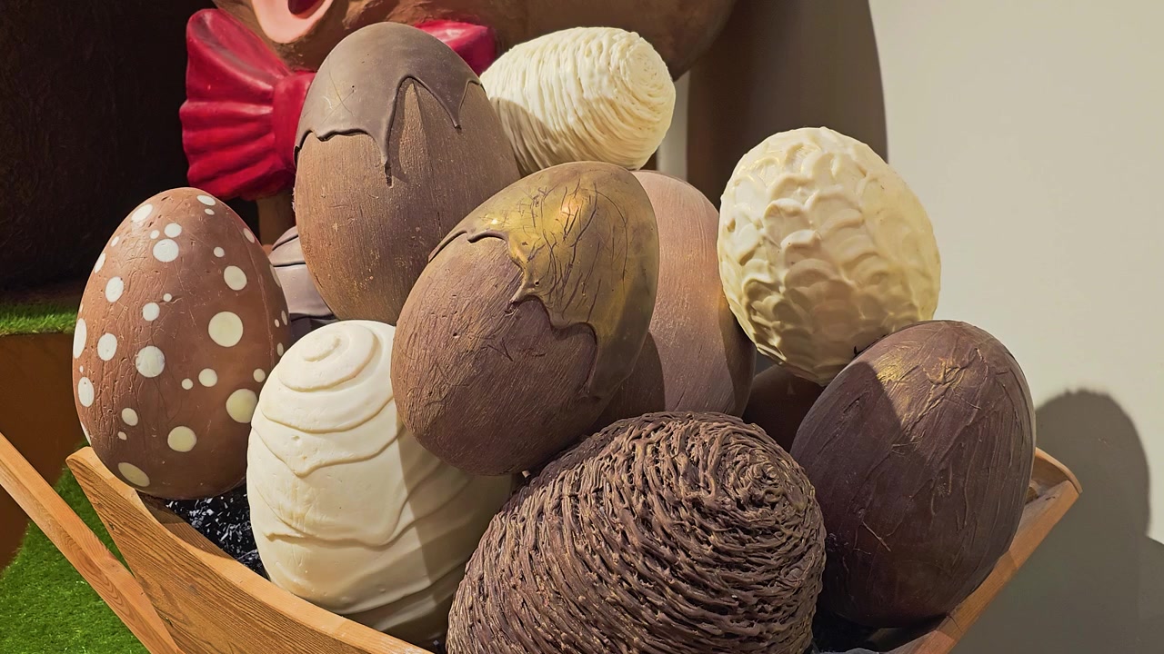 Close up of decorative chocolate Easter eggs in a wooden cart, with textured milk, dark, and white chocolate designs, warm shop lighting, and festive spring holiday display in a candy store.