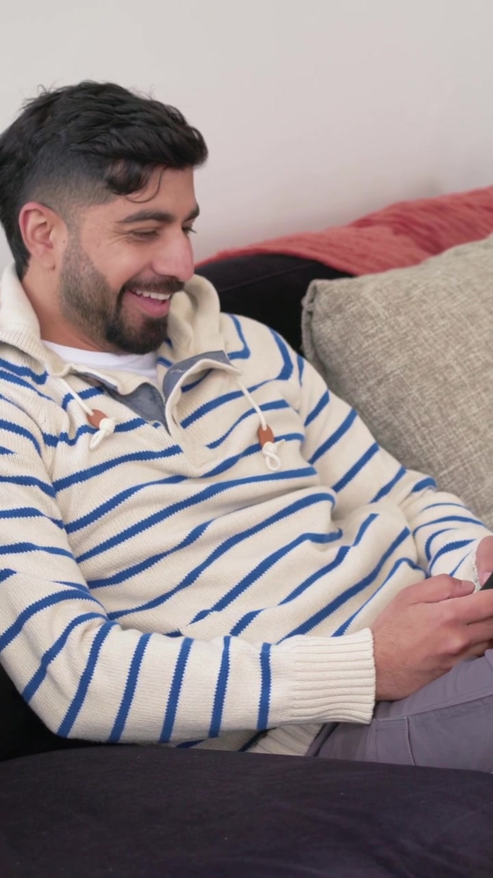 Video verticale: Uomo che si sistema, prende il telefono sul divano di casa, legge messaggi e sorride indossando una felpa con cappuccio.